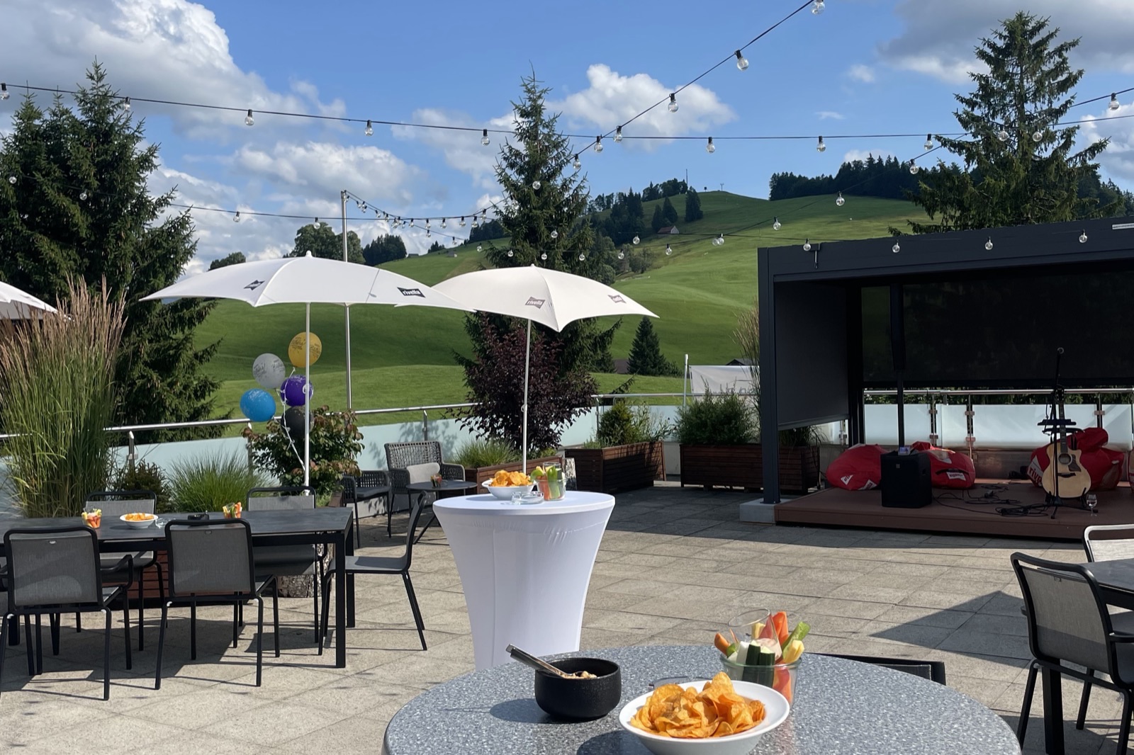Terrasse der Rooftop-Bar im Hotel Allegro: Stehtische und Sonnenschirme unter Lichterketten, dahinter Wiesen und bewaldete Hügel.